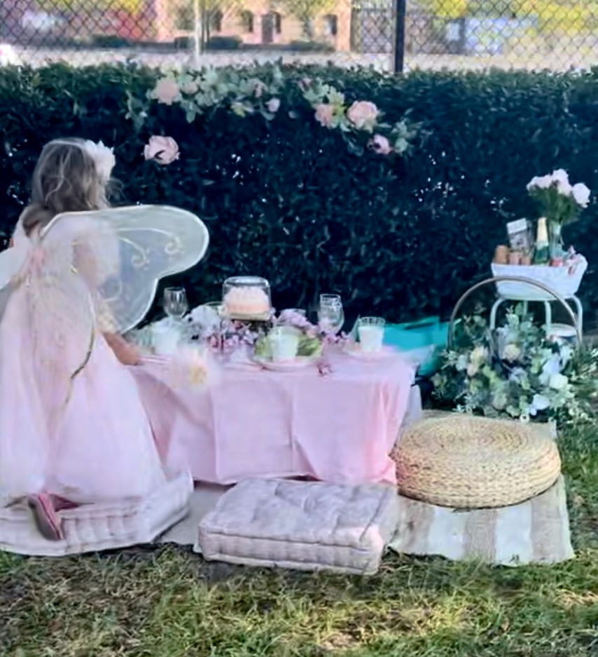 Child in a flower crown at a blush picnic against a hedge pinned with roses.