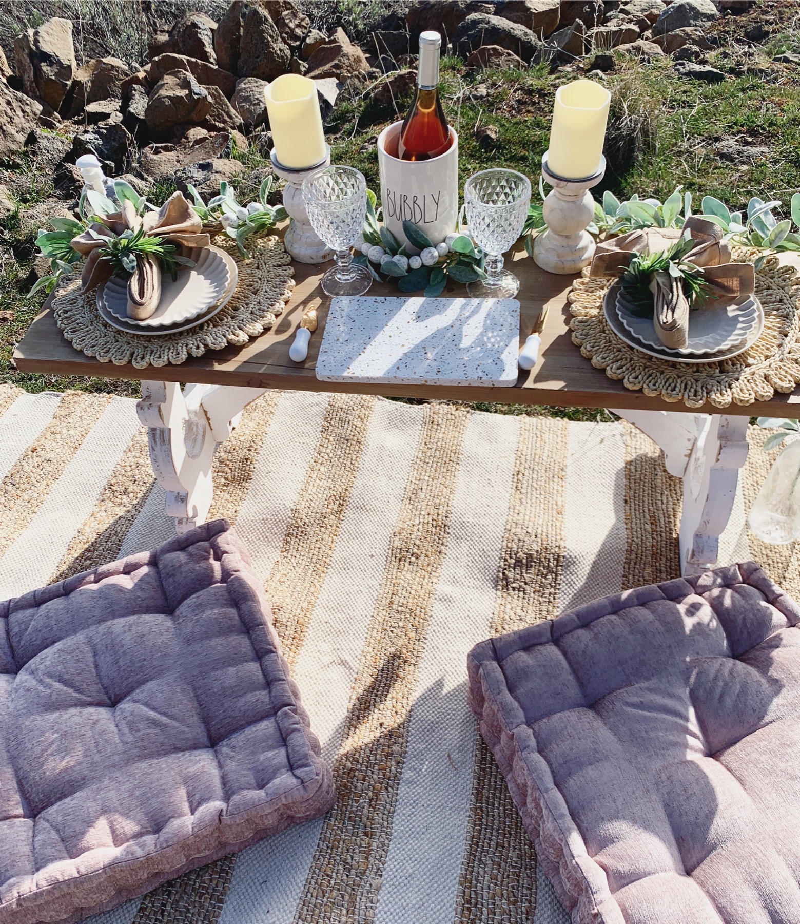 A finished picnic for two: scalloped plates, cut-glass goblets, dusty rose cushions, and a bottle waiting on a striped rug.